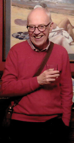 Smiling David Roberts is standing in front of a painting with a drink in his hand.