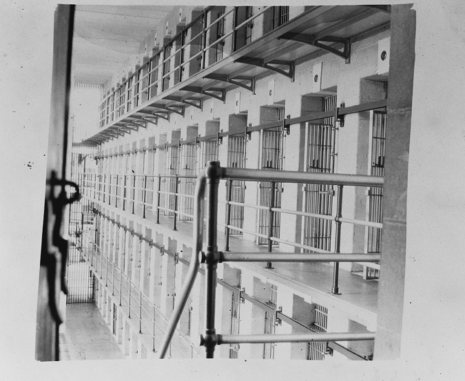 A black and white photograph shows four floors of prisoner cells all facing a central area. Each floor has about 20 cells in a row side by side, with each cell having its own metal barred door. There is a narrow walkway with a metal rail in front of each row of cells that is open to an inside central area of the building.