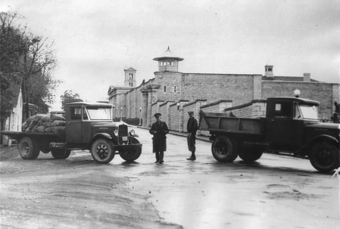 A black and white photograph of two older trucks blocking a street that leads to the front of a large brick building with turrets, which is the penitentiary. One truck has a flatbed piled high with full sacks, and the other truck is a dump truck style. Two men are standing on the street between the two trucks.