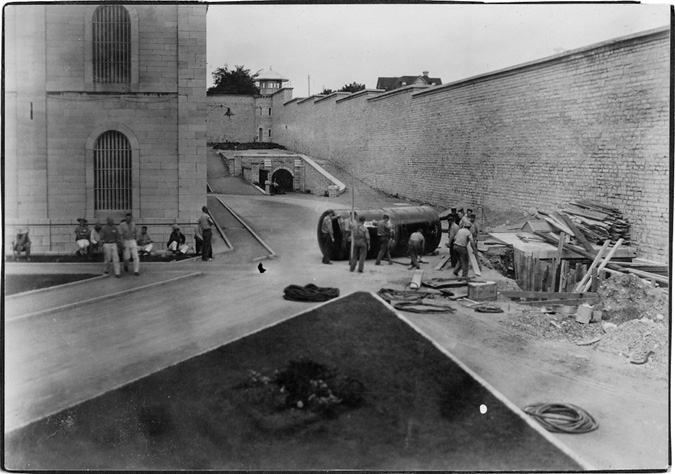 A black and white photograph. On the left is a brick building with pavement surrounding it. On the right is a very large brick wall. A paved area is between the building and the wall. Men are shown taking a break in front of the building with some sitting and some standing. There is a large hole in the pavement next to the wall, and some men are working next the hole with a large black drum behind them.
