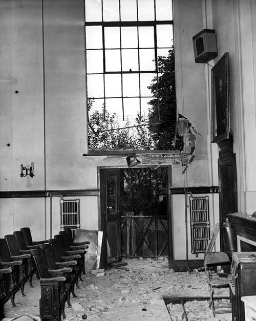 A photograph of the interior of the Labour Temple auditorium. A large window is shattered and the lower wall surrounding it is severely damaged.  Theatre seating is on the left and the floor is covered with glass shards and debris from bomb blast.