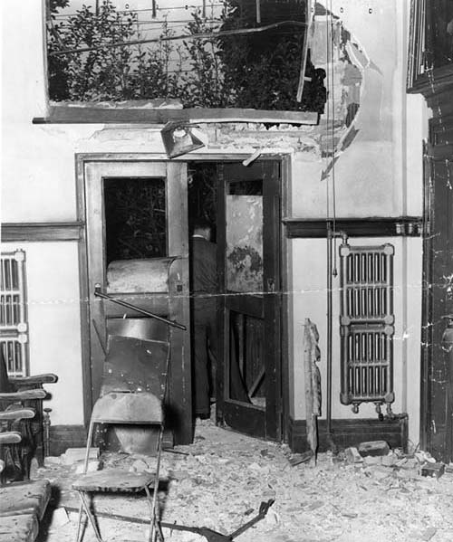 A close-up photograph of the damage done to the Labour Temple auditorium. A large window is shattered and the lower wall surrounding it is severely damaged. The doors beneath the window have broken windows and the wood panel of one of the doors is gone. The floor and chairs are covered with glass shards and debris from bomb blast.