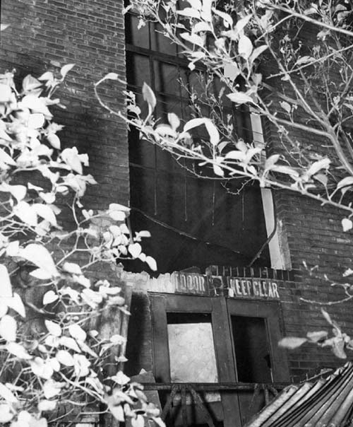 A photograph of the exterior of the Labour Temple after the bombing.  There is severe damage to the window and the doors beneath.  Some of the brick has been damaged and pieces are missing.