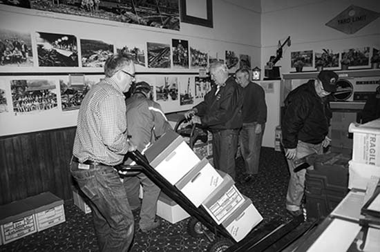 A photograph with 5 men in a room that has various large photos on the walls.  One man has a dolly with 4 boxes that he is moving.  There are various other boxes on the floor.