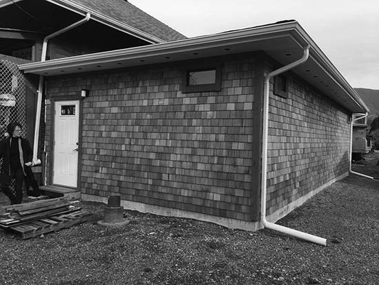 A photograph of the outside portion of a building.  The exterior looks to be finished with shingles and there is a pile of wood on the ground in front of the door.
