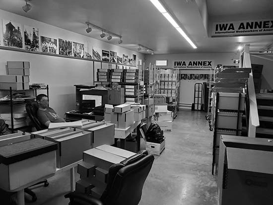 A photograph of the inside of a room.  The walls have numerous photos. There are two banners up high that say, IWA ANNEX with the wording International Woodworkers of America underneath.The room is full of boxes and metal carts. There is a man sitting in an office chair.