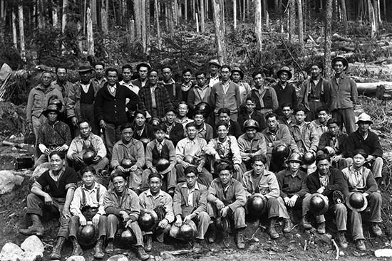 A group photo of 40 to 50 men. They are arranged in 4 rows with the first two rows sitting and the back two rows standing. Many of the men are holding hard hats. Almost all of the men in the photograph appear to be Asian.