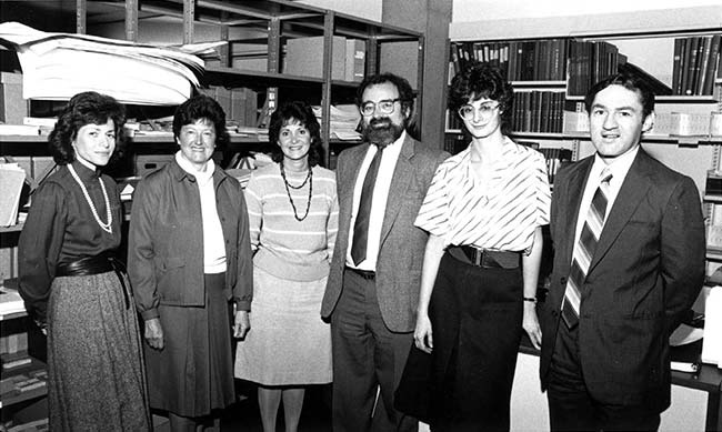 A photograph with 4 women and 2 men standing and posing in front of bookshelves in a room. 