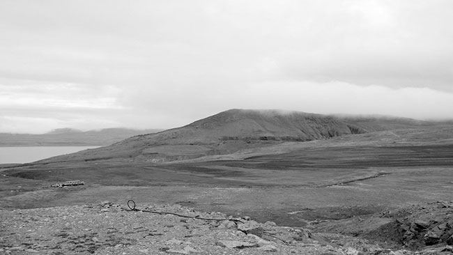 A 2011 photograph of a now-vacated plain that was Nanisivik townsite. In the distance, a rock shelf can be seen through fog and a body of water partially glimpsed.