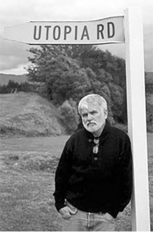 A photograph of Mike Davis wearing a dark shirt and jeans posing beneath a street sign with the words Utopia Road written on it.