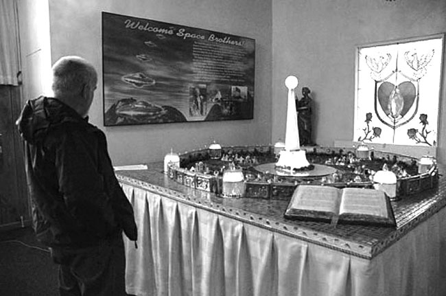 A photograph of Mike Davis observing a diorama of a Tesla Tower at the Church of Unarius. A bible sits open in the foreground and on the wall 2 framed religious pictures hang. 