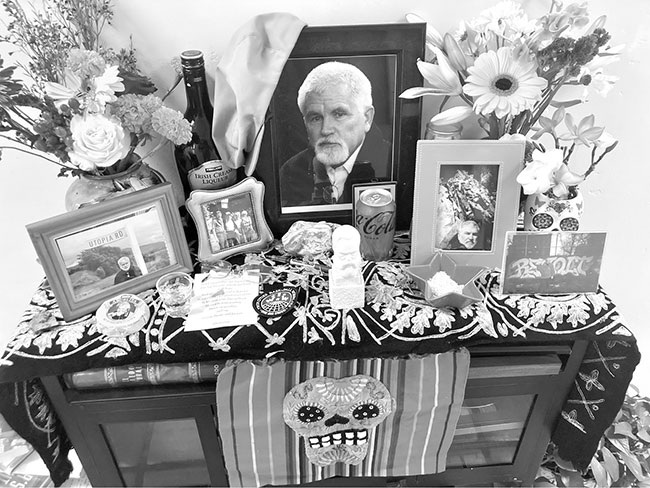 A photograph of a table-top spread with objects memorializing Mike Davis. The spread consists of a large picture of Davis in the centre of the frame, several other pictures of Davis displayed on either side, a tablecloth with a Day of the Dead face on it, some flowers, mementos, and a bottle of liquor.