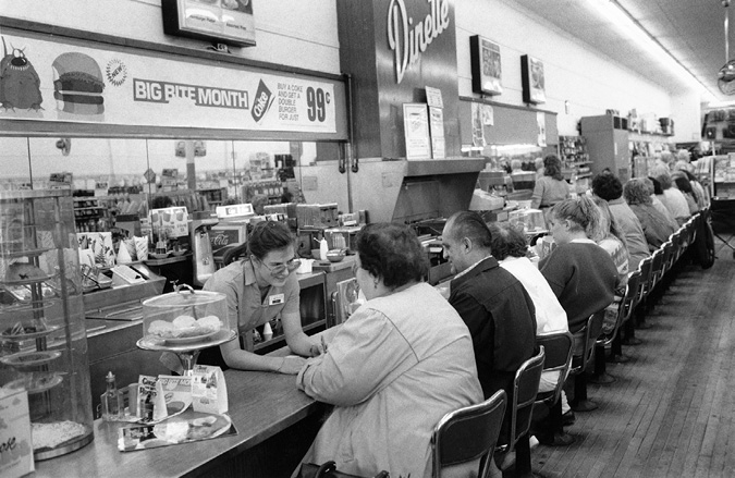 A black and white photograph of a 1970's style lunch counter at a dinette. The photo was taken at an angle, showing about 11 patrons sitting at the counter. There is a female server leaning over the counter speaking with a customer.