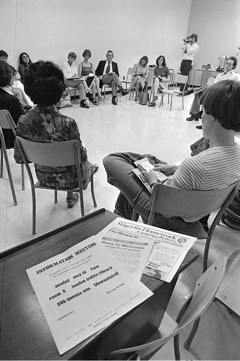 A black and white photo of a gathering of people in a large room. There are between 10-15 people sitting in chairs in a circle. There are papers on a table in the room. One has the headline "Wages for Housework".  Another says "Money for Waitresses" and a leaflet  saying "Information meeting, Sunday May 15, 2 pm, room 3, London Public library.