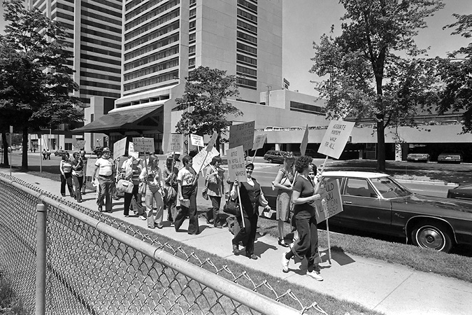 A black and white photo of a protest in a downtown area. The photo shows about 20 men and women walking in a line on a sidewalk with signs referencing minimum wage.