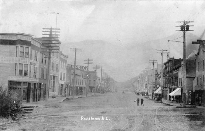 A black and white photo of street-scape in Rossland, B.C.. The road is made of dirt and there is a wagon parked on the side.  The sidewalks are made of wood.  