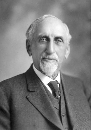A head shot of an older white man wearing a suit.  He is partially bald and has a mustache and goatee.