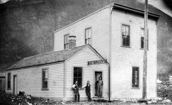 A photograph of a building with 3 men standing outside.  Two are leaning beside a window and one stands int he doorway.  There is a sign above the door that says "The Ledge"