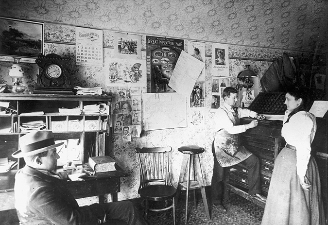 A photograph of a room with 3 people.  One man with a hat sits while one man wearing an apron is standing at a printing press, and a woman is standing next to him.  