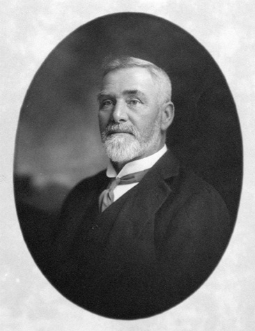 A head shot of an older white man wearing a suit.  He has a gray mustache and beard.