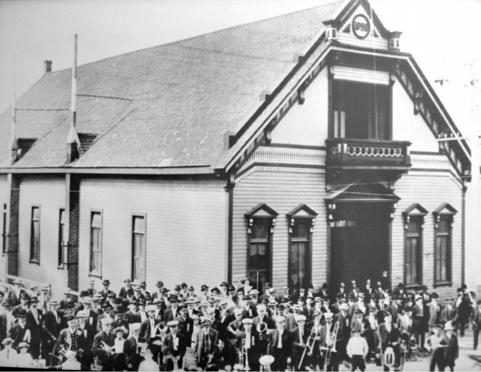 A photograph of a building with a band standing outside. The building has a balcony. The band has  75 to 100 members and they are holding their instruments.