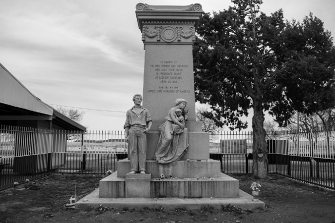 A photograph of a large white monument. The center pillar says "In memory of the men, women, and children, who lost their lives in freedom's cause at Ludlow, Colorado, April 20, 1914.  Erected by the United Mine Workers of America". At the front of the monument is a statue of a man with his hand resting on his waist, and next to him is a statue of a woman holding a child.