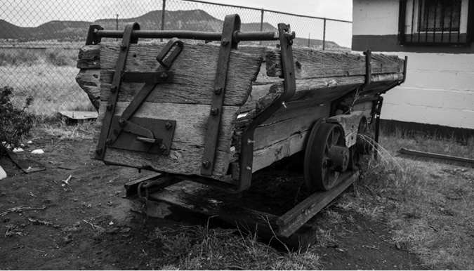 A photograph of an old mine-cart made of wood and iron.  Its wheels are on a section of  railway track.