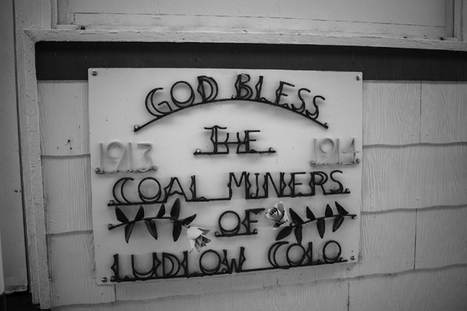 A photograph of a sign on the side of a building. The words are made of iron and say, "God bless the coal miners of Ludlow, Colorado".  The years 1913 and 1914 are on either side of the sign. 