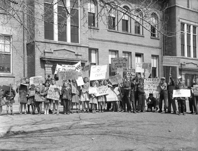 A photograph of a group of  about 30 children holding placards and protesting the rise in price of candy bars. They are standing in front of a school.