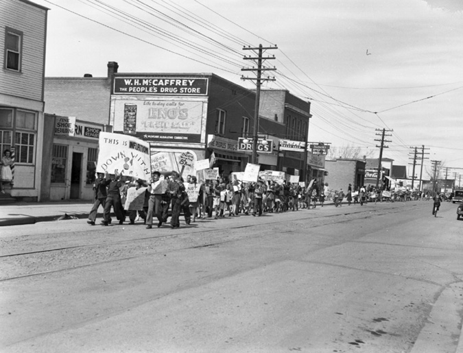 A photograph of a group of children carrying placards and protesting the rise in price of candy bars. They are walking down a business street in protest.