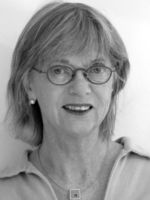 A grayscale portrait of a woman with shoulder-length hair wearing glasses and a light-colored collared shirt. She is smiling slightly and wearing a square pendant necklace and pearl earrings.