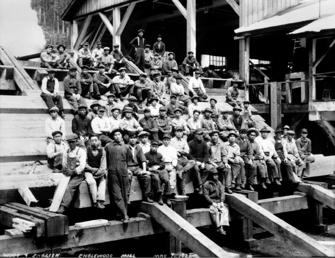 Dozens of Asian immigrant workers and other sawmill workers sit in rows in front of a mill, looking toward the camera in a posed photograph. The outside of the mill, as well as the forest behind it, can be seen in the background.