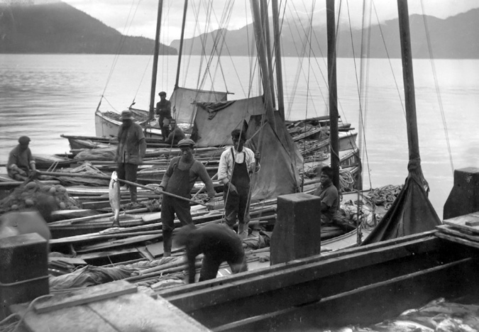 Eight Japanese Canadian fishermen stand on a set of small, docked fishing boats that are filled with salmon. One worker in the middle ground lifts a salmon that is speared on a stick. In the foreground, a large container filled with salmon is visible. In the background, the water extends toward forested mountains.