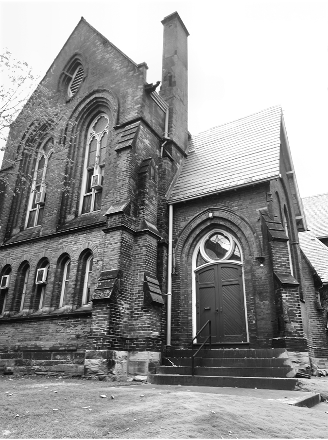 A black-and-white photo of a historic brick church with Gothic architectural features. The building has tall, narrow arched windows with decorative trim and air conditioning units installed in several of them. A large arched doorway with double wooden doors and a semicircular window above is visible to the right, accessed by a small set of stairs with a railing. The brickwork shows signs of age and weathering, and a tall chimney rises from the roof.