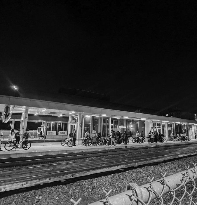 A black-and-white nighttime photo of UP Express Bloor station A group of people, many with bicycles, are gathered along the platform, standing or sitting near the building. Train tracks run in the foreground The dark sky above contrasts with the well-lit station, giving a sense of quiet anticipation or waiting.