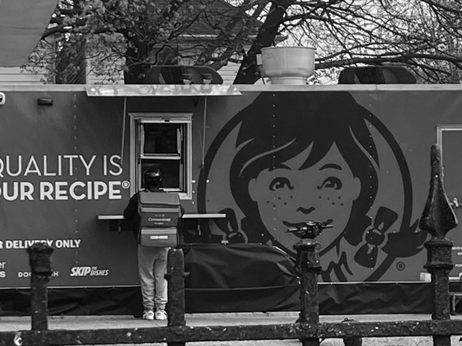 A photo of a food truck featuring the large logo of Wendy's, a fast-food restaurant chain. A delivery person wearing a helmet and carrying a large insulated delivery backpack is standing at the service window of the truck, seemingly picking up an order.