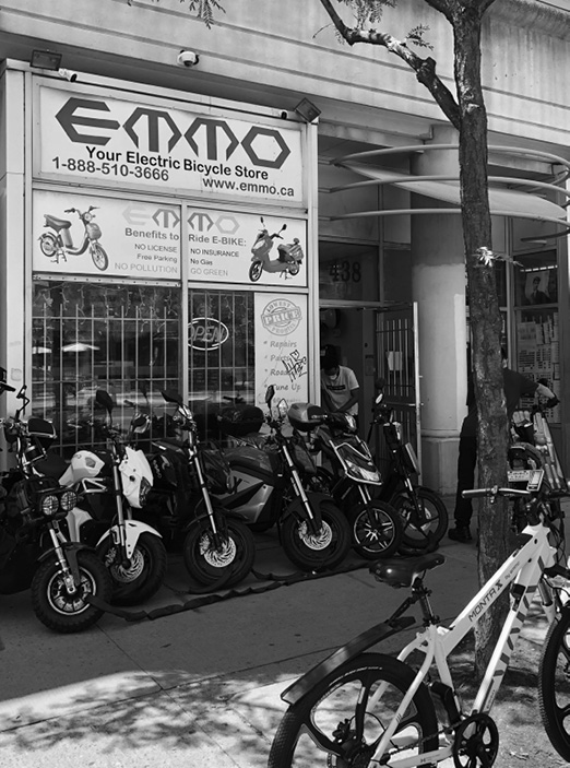 A photo of the storefront of EMMO, an electric bicycle store. The sign above the shop displays the store's name along with benefits of riding an e-bike such as no license, no insurance, free parking, and no pollution. In front of the shop, a row of various electric bikes and scooters is tightly lined up on the sidewalk.