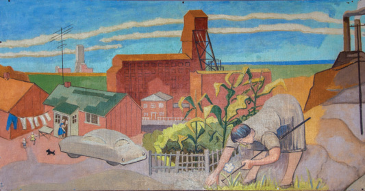 The first section of a painted mural scene depicting a stylized industrial town with a mix of domestic and industrial elements. In the foreground on the right, a woman with dark hair bends over a garden, tending to plants with a gardening tool in hand. Behind her, tall corn stalks grow along a fence. To the left, modest red houses line the street; one woman stands at the door of a green-roofed house, watching two children and a small black dog. A vintage car is parked in the driveway. Further back, large red-brick industrial buildings dominate the skyline, with a mine headframe structure rising above them. Smokestacks and a flat landscape stretch into the distance under a vivid blue sky with layered clouds. The overall composition blends scenes of labor, home life, and industry, evoking a working-class community.